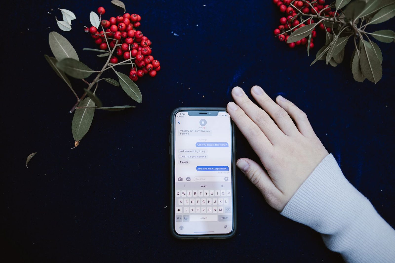 Mano appoggiata accanto a uno smartphone che mostra una conversazione di messaggistica, su sfondo blu con rami dalle bacche rosse