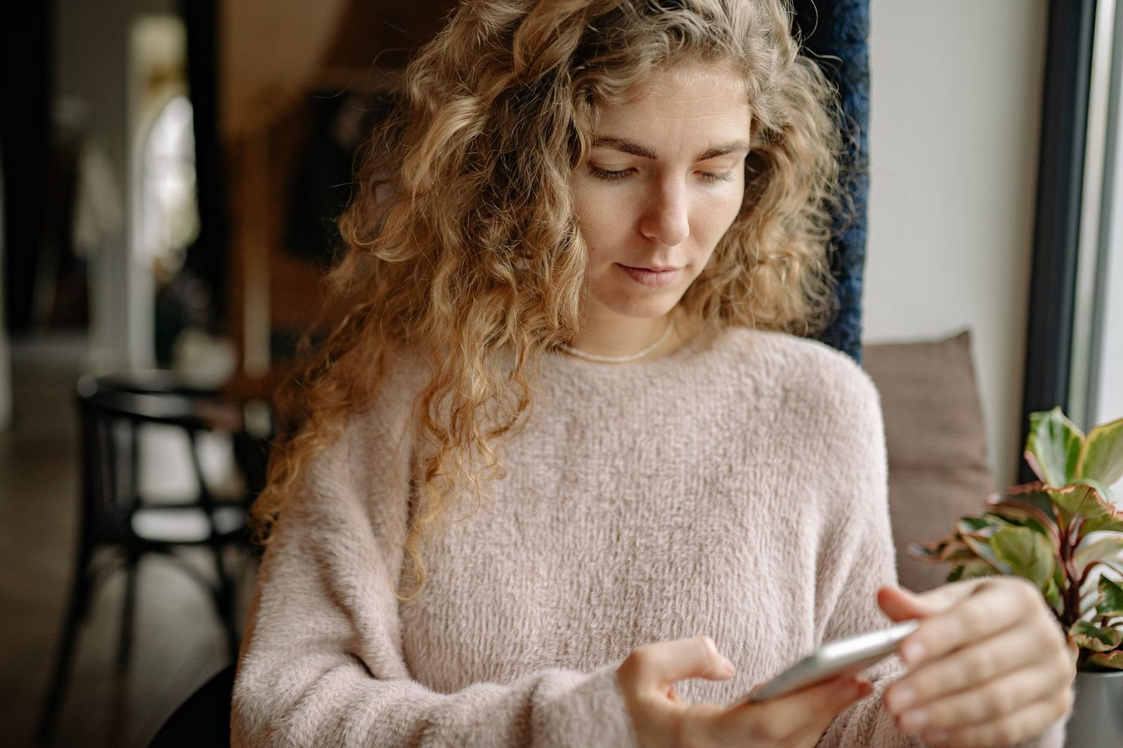 Jonge vrouw met krullend haar die bij een raam zit en een bericht op haar smartphone bekijkt