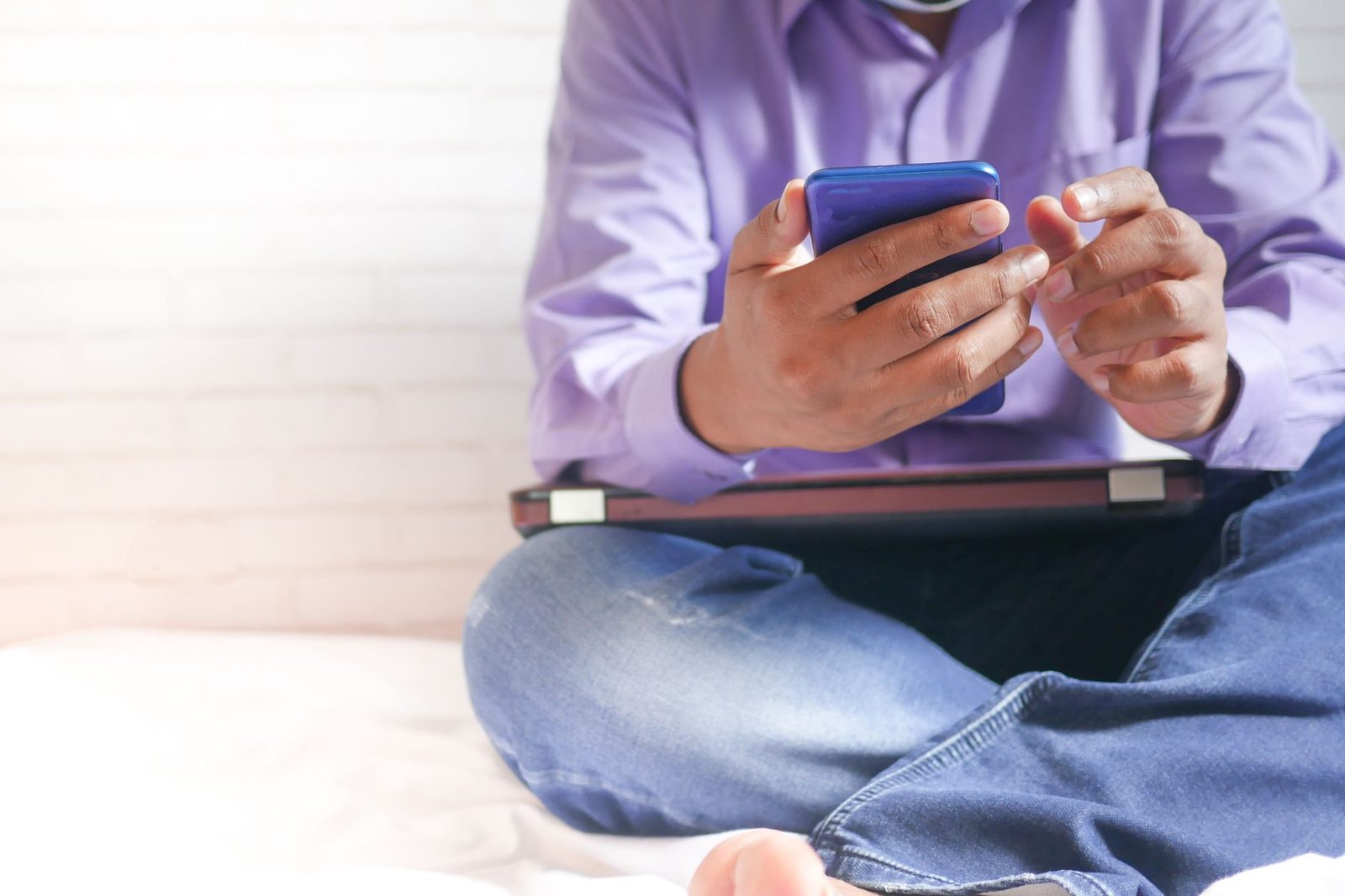 Man die met gekruiste benen op een bed zit in een paars overhemd, zijn blauwe smartphone bekijkt met een laptop op schoot