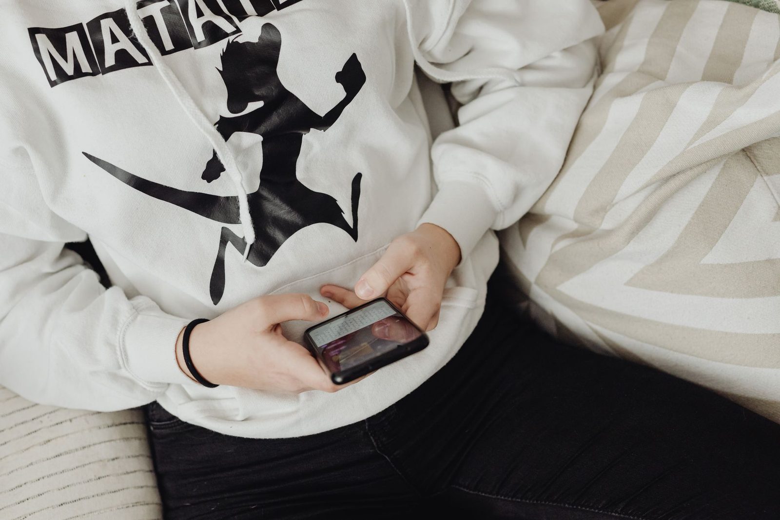 Persoon in een witte hoodie die zittend berichten bekijkt op een smartphone die hij met twee handen vasthoudt