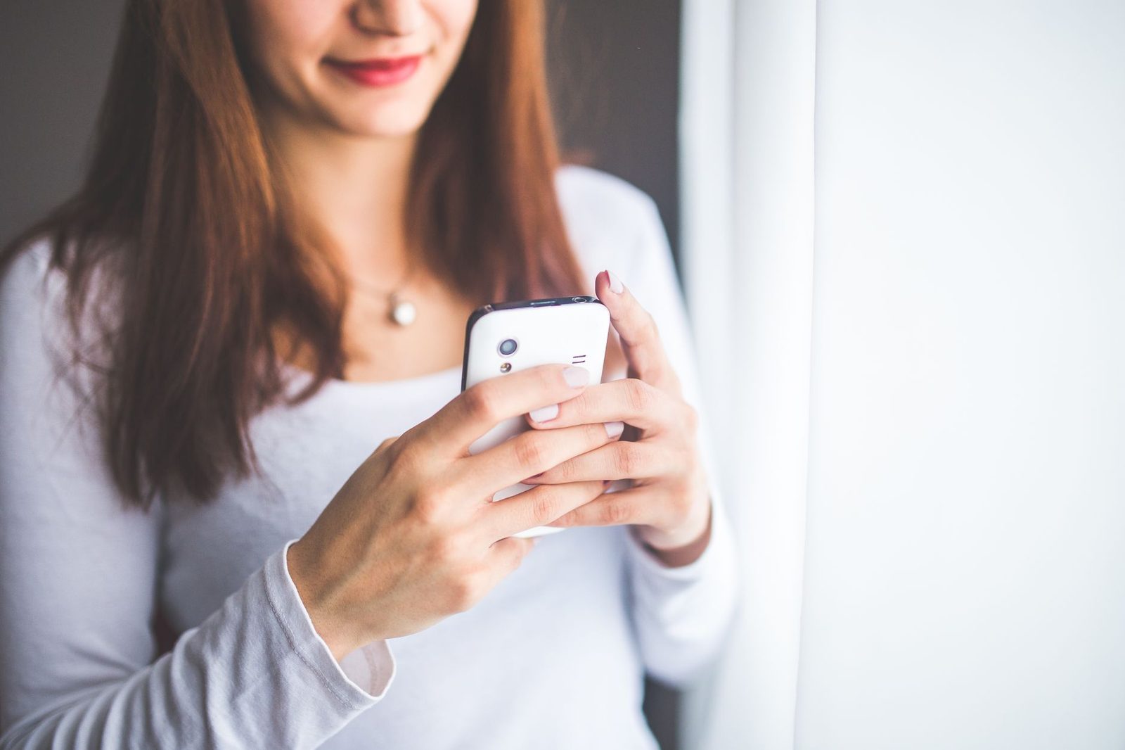 Uśmiechnięta młoda kobieta w białej bluzce trzymająca smartfon w obu dłoniach przy oknie