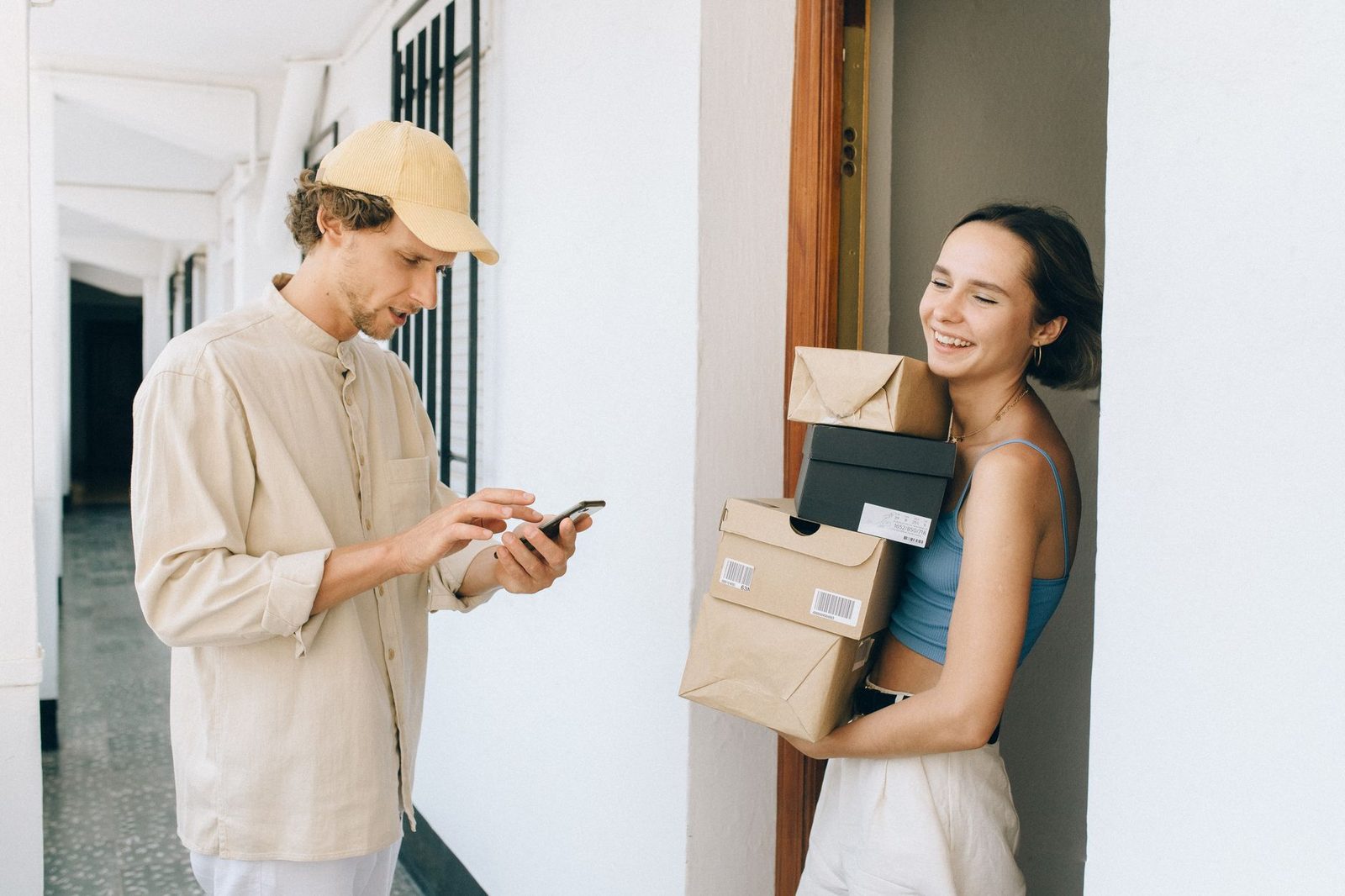 Corriere che consulta lo smartphone davanti a una giovane donna sorridente che tiene vari pacchi in un corridoio