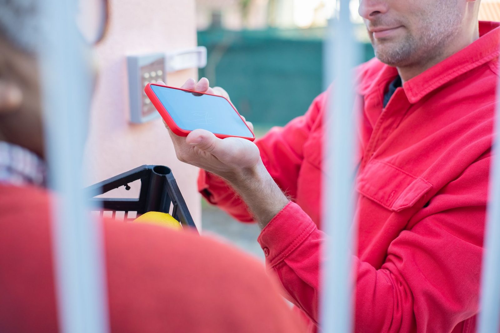 Uomo con giacca rossa che tiene uno smartphone davanti a un cancello, accanto a una cassa di consegna