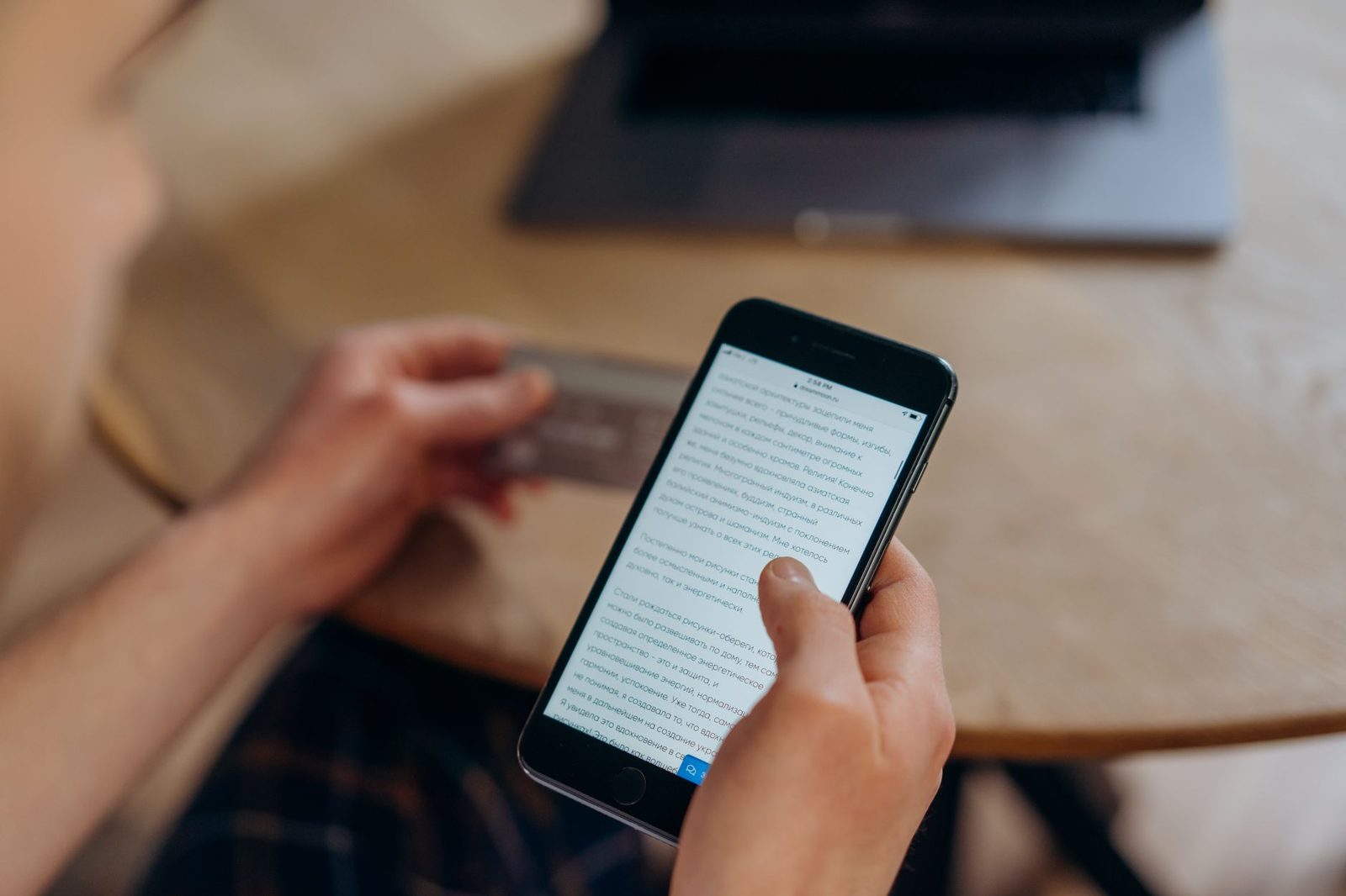 Persona sosteniendo un smartphone y una tarjeta bancaria, leyendo un texto en la pantalla frente a un ordenador portátil