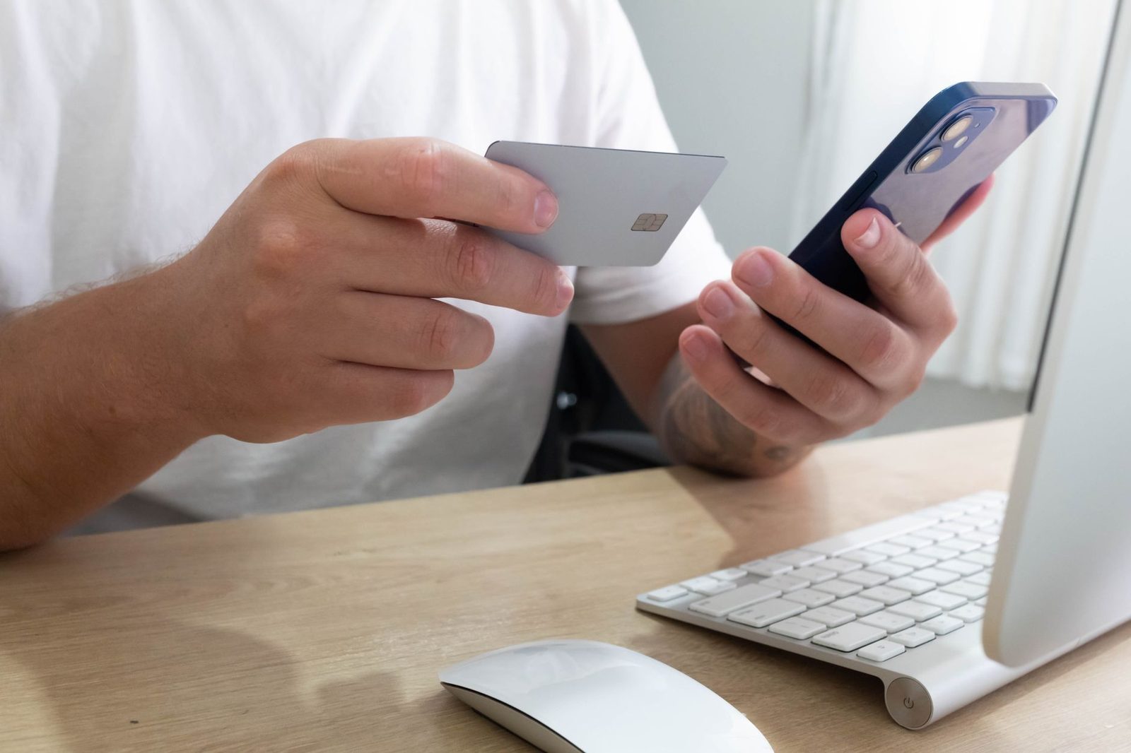 Hombre sosteniendo una tarjeta bancaria y un smartphone frente a un ordenador sobre un escritorio de madera