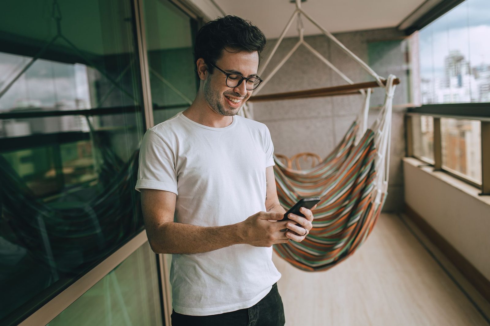 Uomo sorridente con occhiali che guarda lo smartphone su un balcone, vicino a un'amaca sospesa