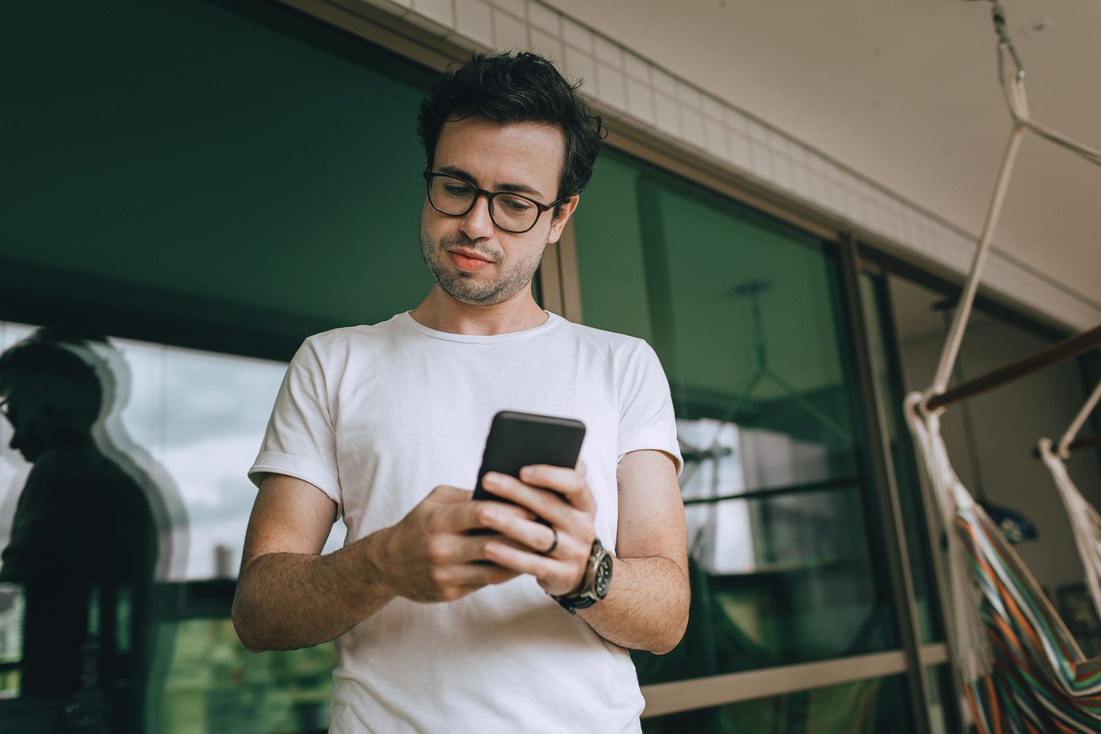 Uomo con occhiali e maglietta bianca che guarda un messaggio sul cellulare con aria preoccupata