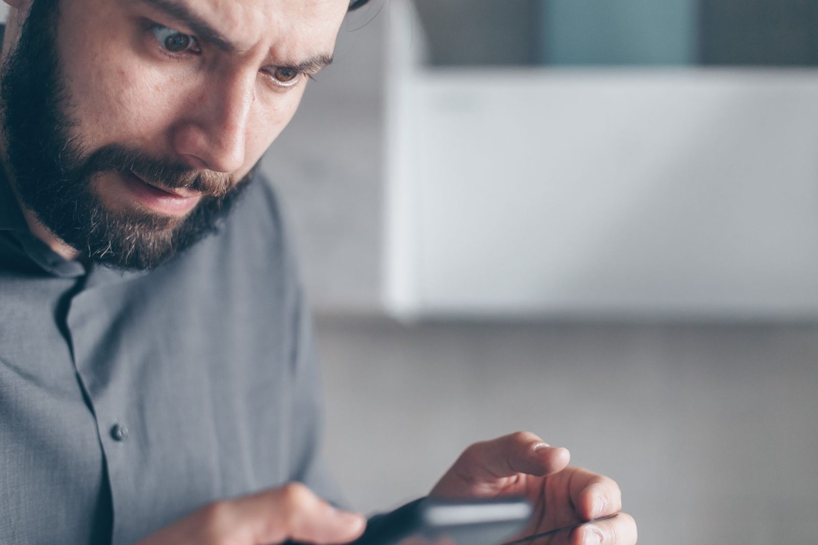Uomo con la barba dall'aria preoccupata che guarda lo schermo dello smartphone tenendolo con entrambe le mani
