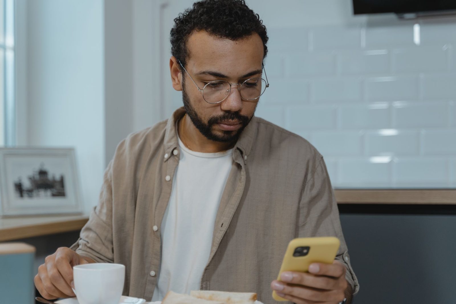 Homme à lunettes lisant un message sur son smartphone jaune devant un café et des tartines