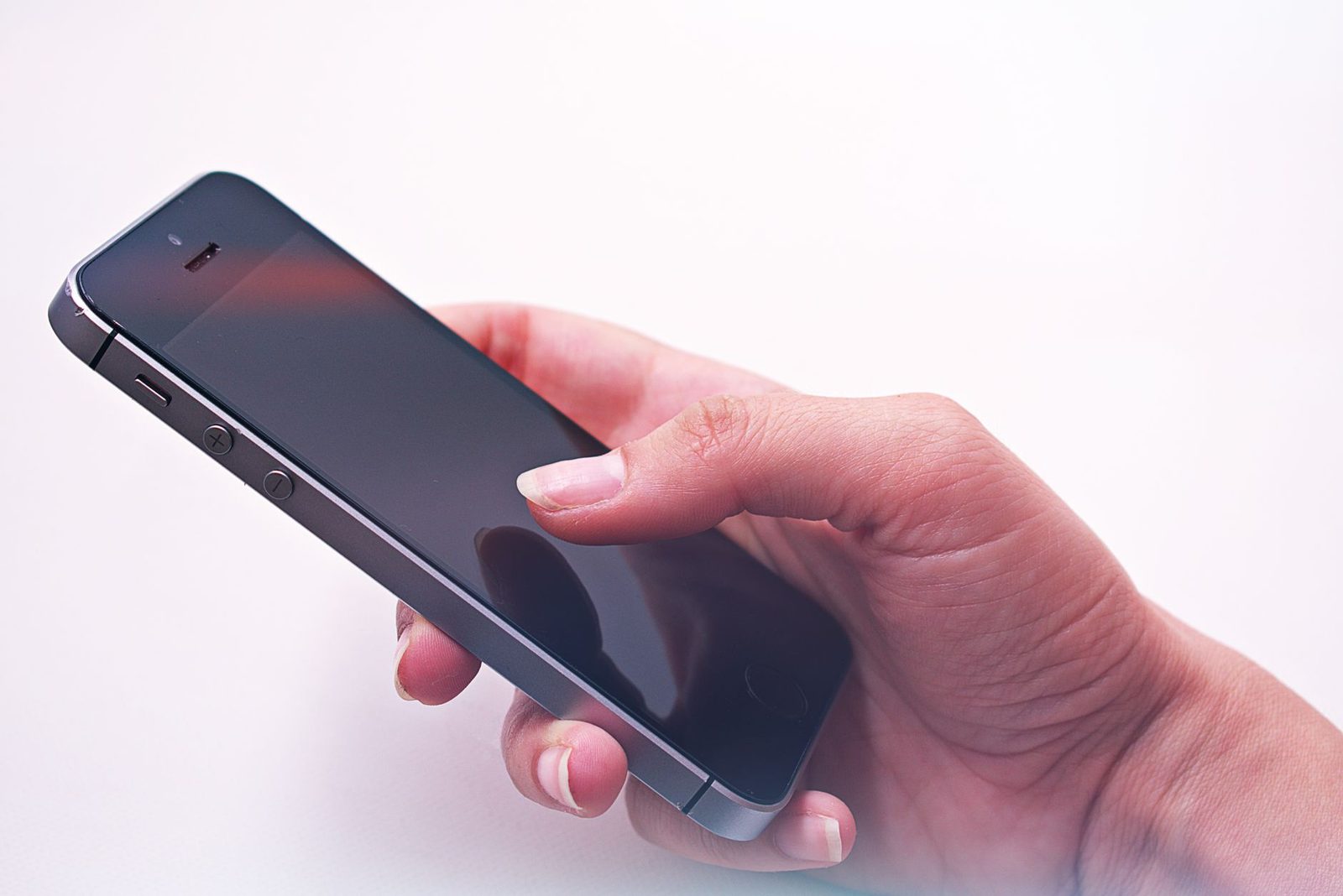 Mano che tiene uno smartphone nero con lo schermo spento, il pollice appoggiato sul display, su sfondo bianco