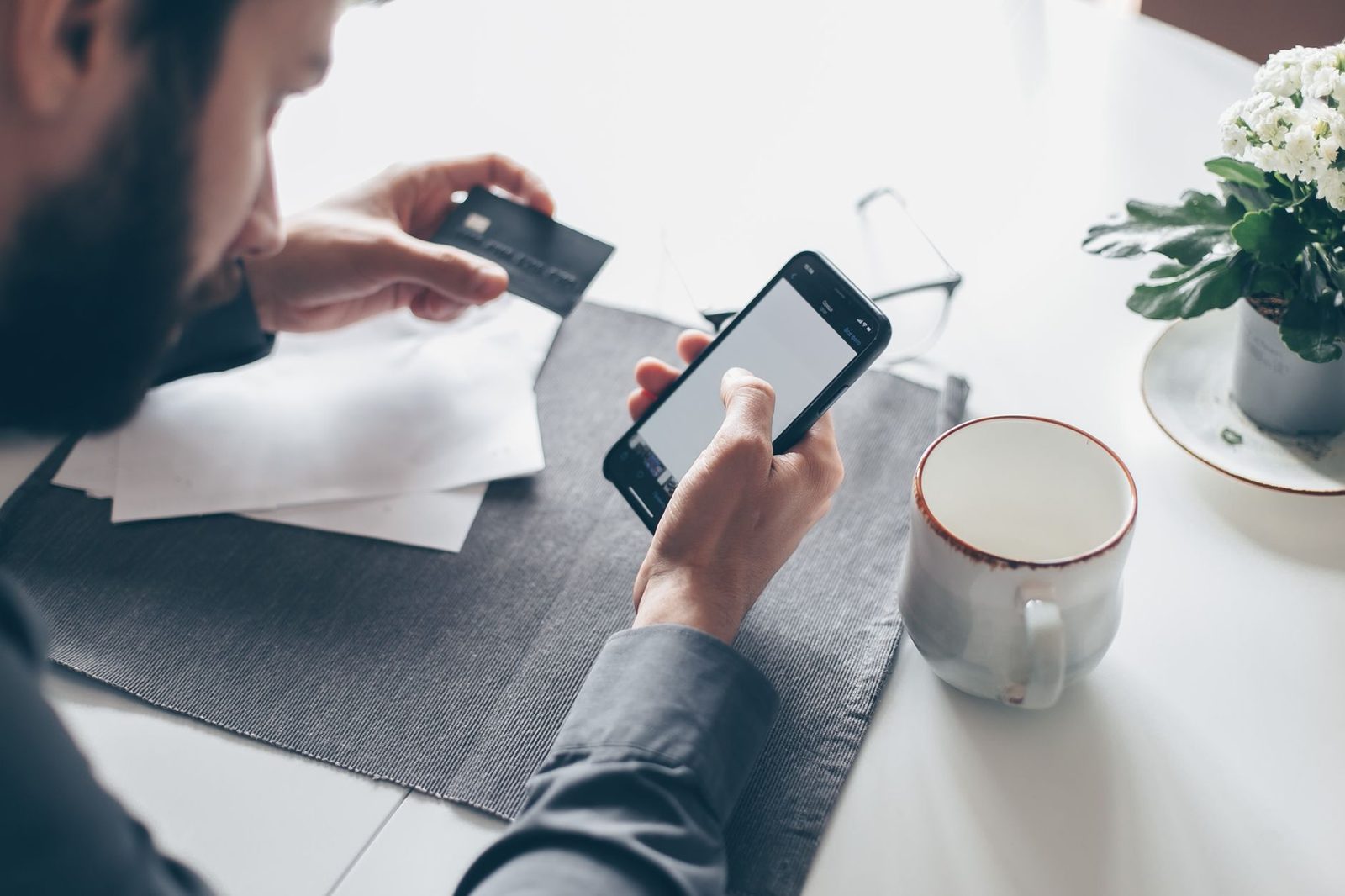 Mann mit Bankkarte in der Hand schaut auf sein Smartphone, sitzt an einem Tisch mit einer Tasse