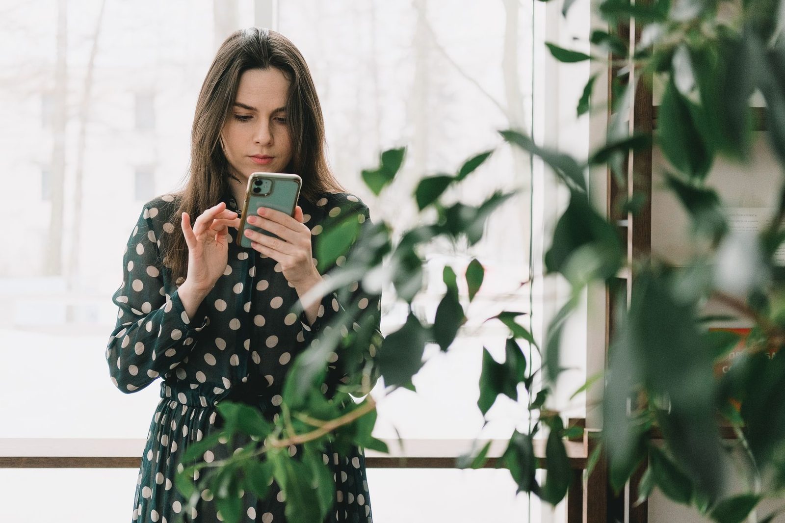 Młoda kobieta w sukience w grochy przegląda smartfon przy oknie, na pierwszym planie zielone liście