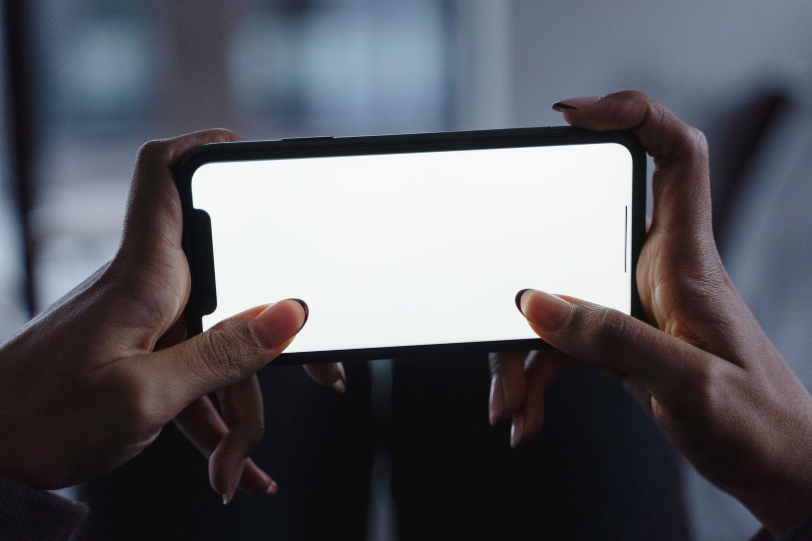 Mani che tengono uno smartphone con lo schermo bianco vuoto in modalità orizzontale, in una stanza buia