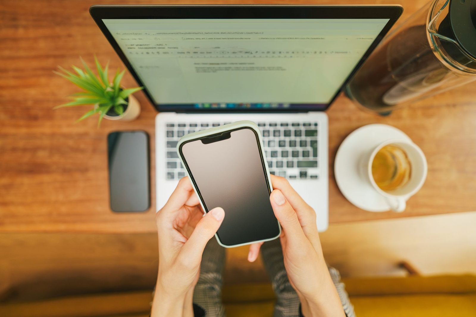 Femme tenant un smartphone à l'écran éteint devant un ordinateur portable, vue de dessus sur un bureau