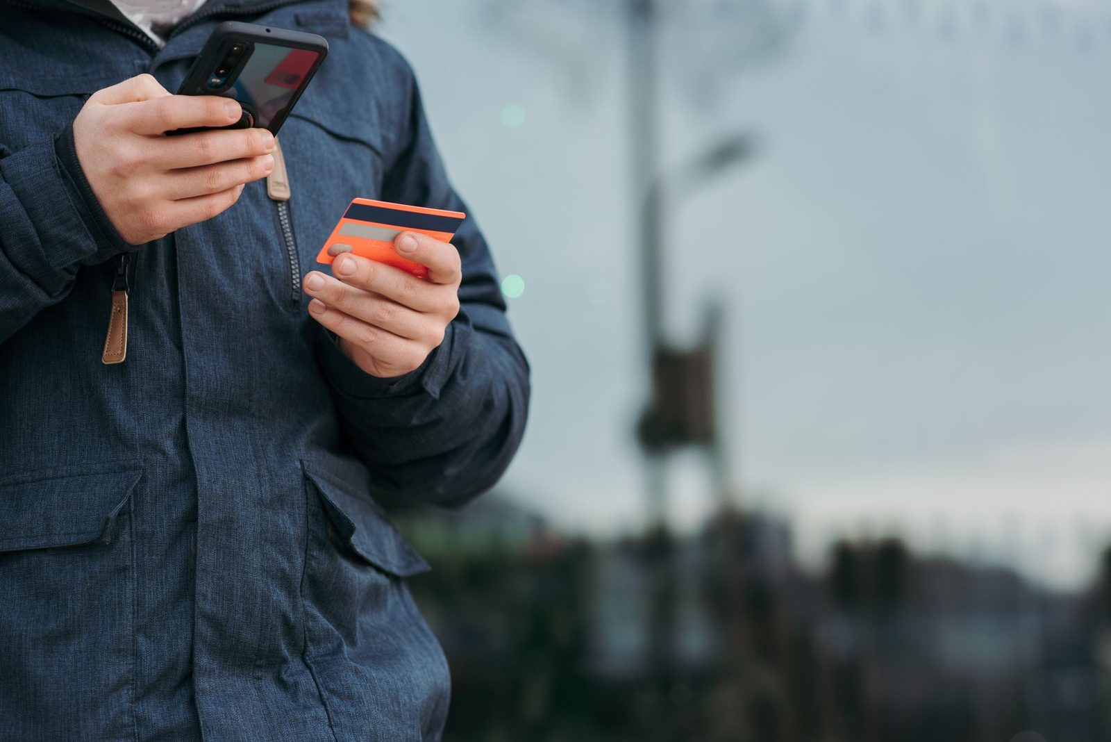 Personne en veste bleue tenant un smartphone et une carte bancaire orange en extérieur