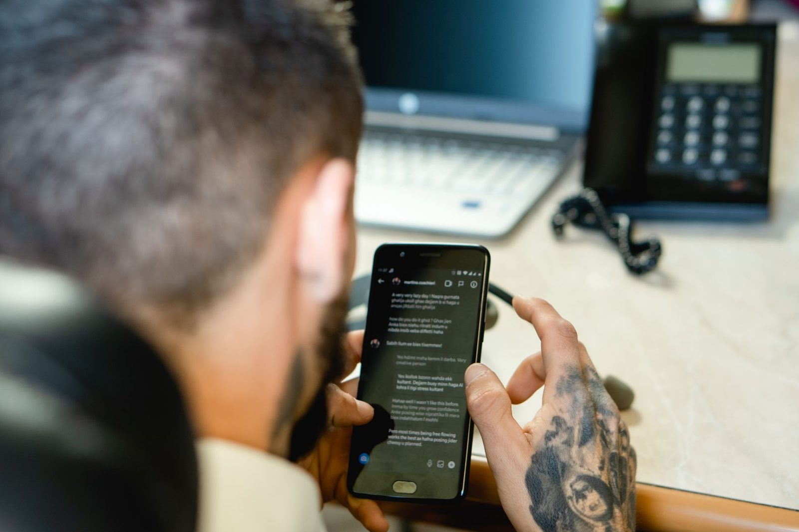 Tattooed man reading messages on his smartphone, sitting at a desk with a laptop and a landline phone