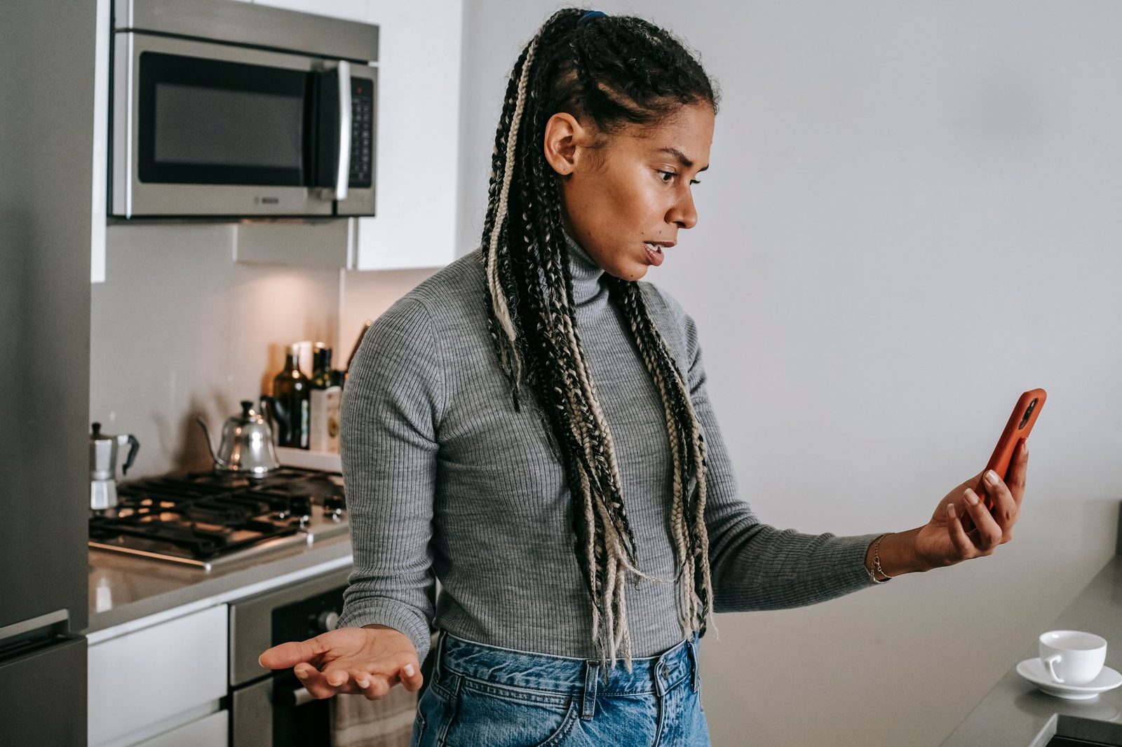 Femme perplexe dans une cuisine regardant l'écran de son smartphone, une main levée en signe d'incompréhension