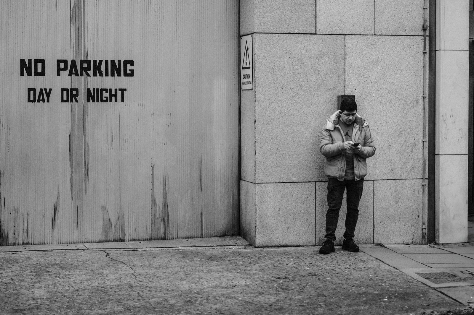 Photo en noir et blanc d'un homme adossé à un mur dans la rue, consultant son téléphone portable