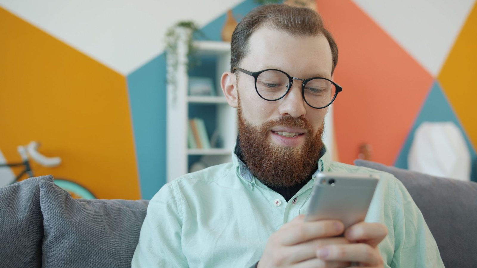 Bearded man in glasses sitting on a sofa, smiling as he reads a message on his smartphone