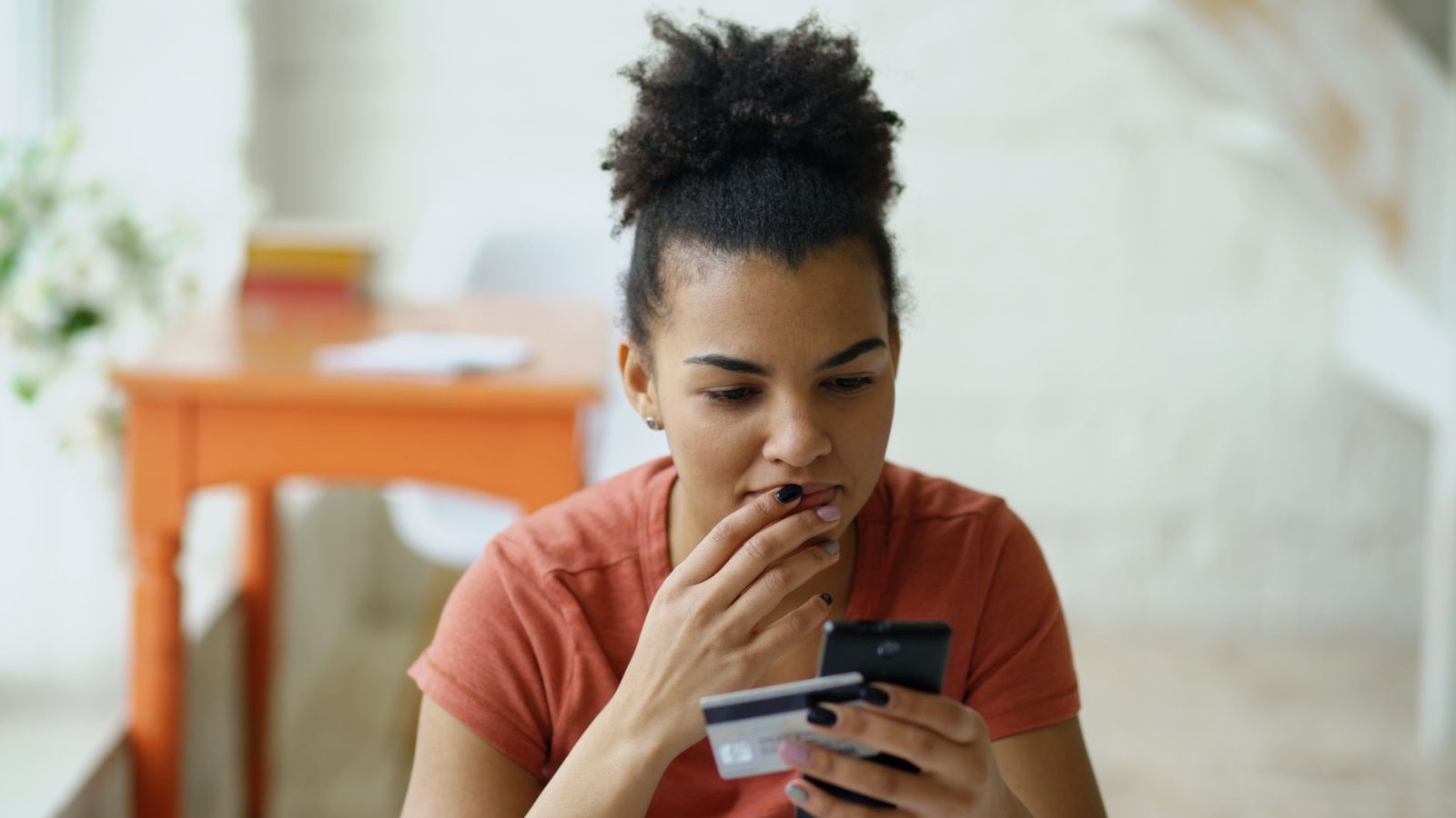 Jeune femme inquiète, la main devant la bouche, regardant son smartphone en tenant une carte bancaire
