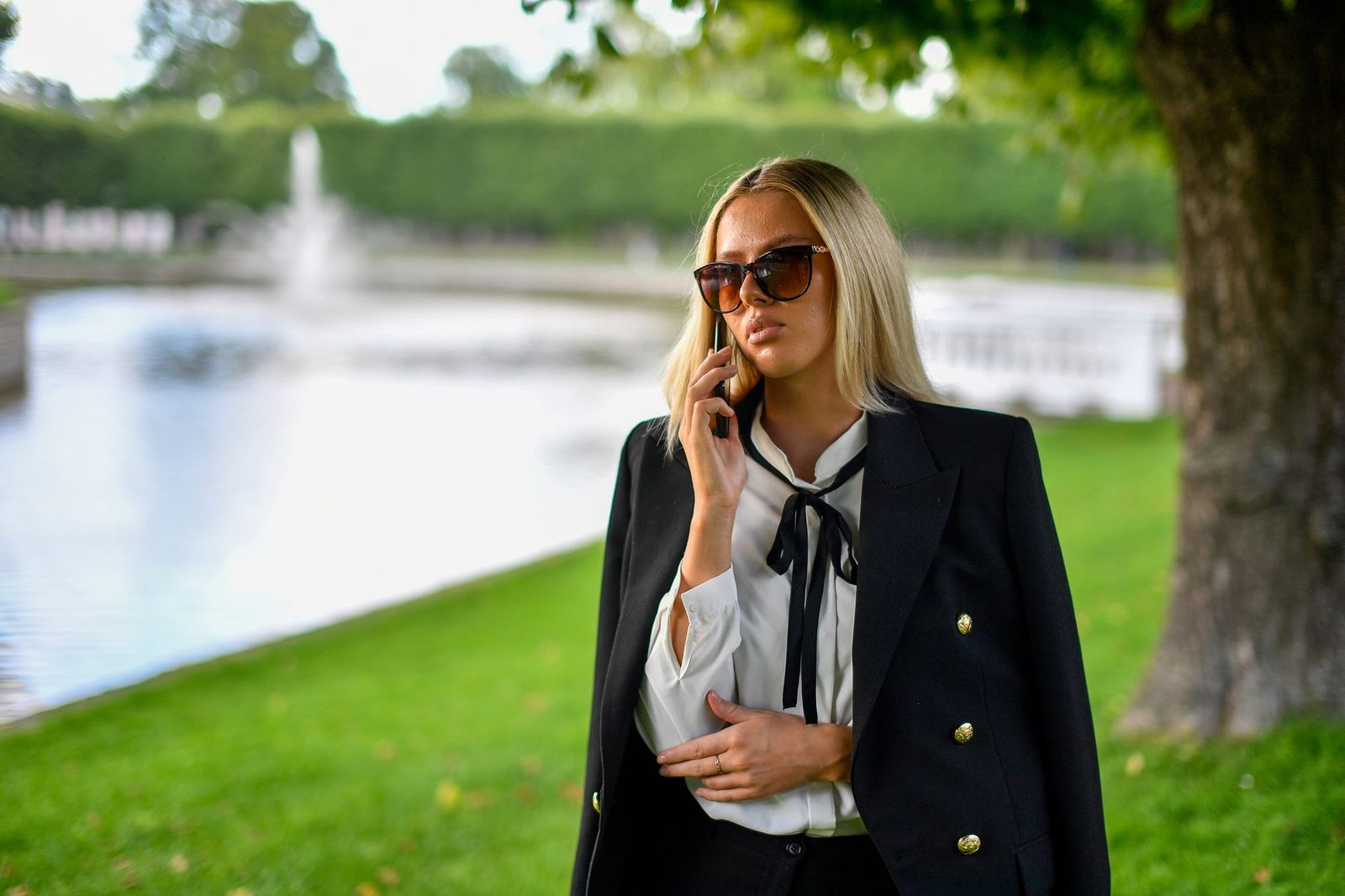 Young blonde woman in a black jacket and sunglasses talking on her smartphone in a park