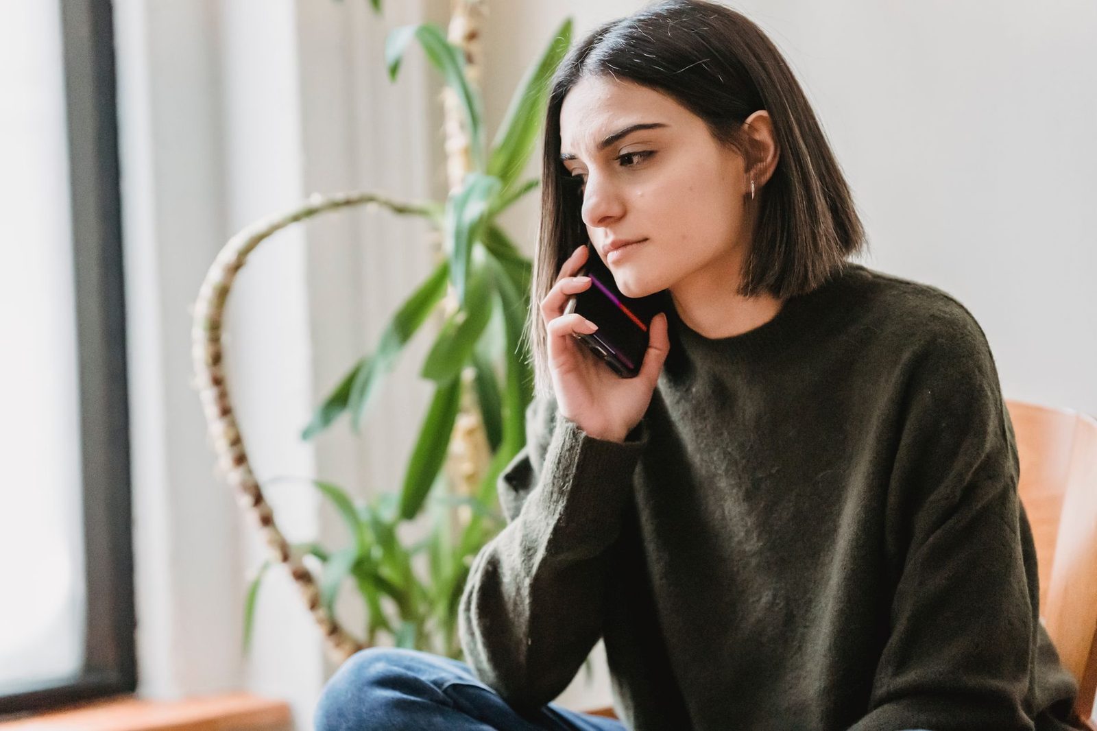 Jeune femme en pull vert, l'air inquiet, téléphone portable à l'oreille, assise près d'une plante verte