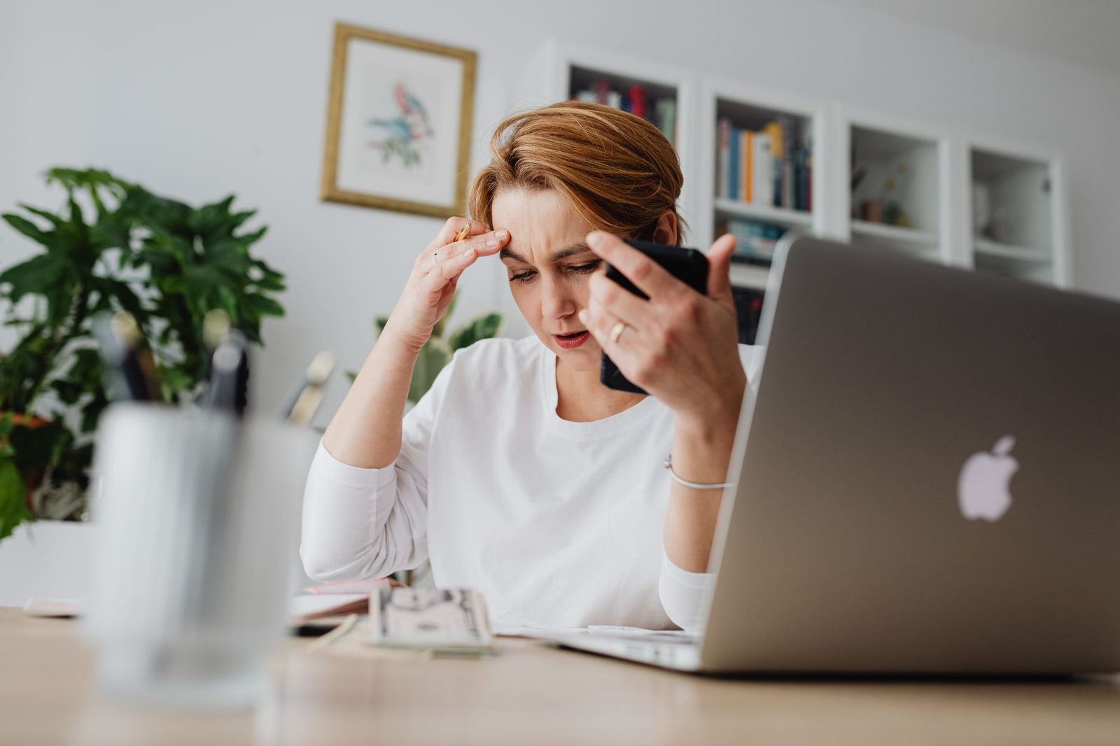 Femme inquiète tenant son smartphone devant un ordinateur portable, la main sur le front