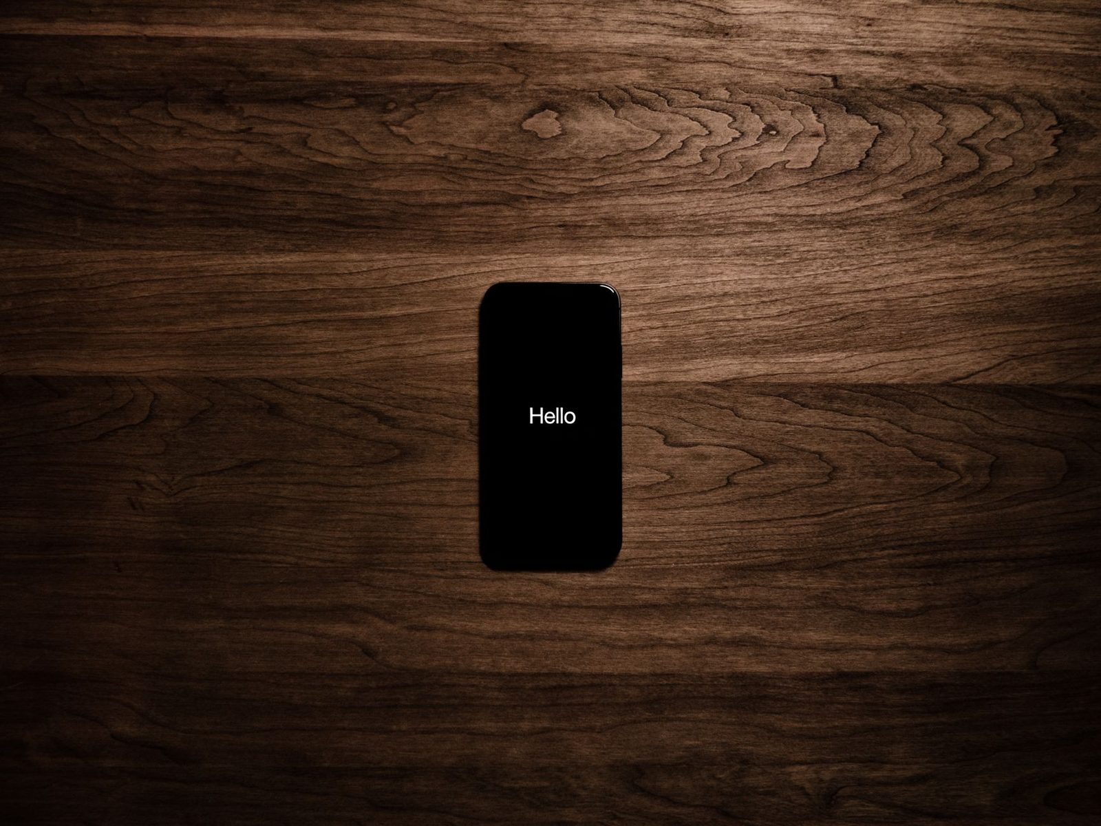 Smartphone posé sur une table en bois sombre, écran noir affichant le mot « Hello »