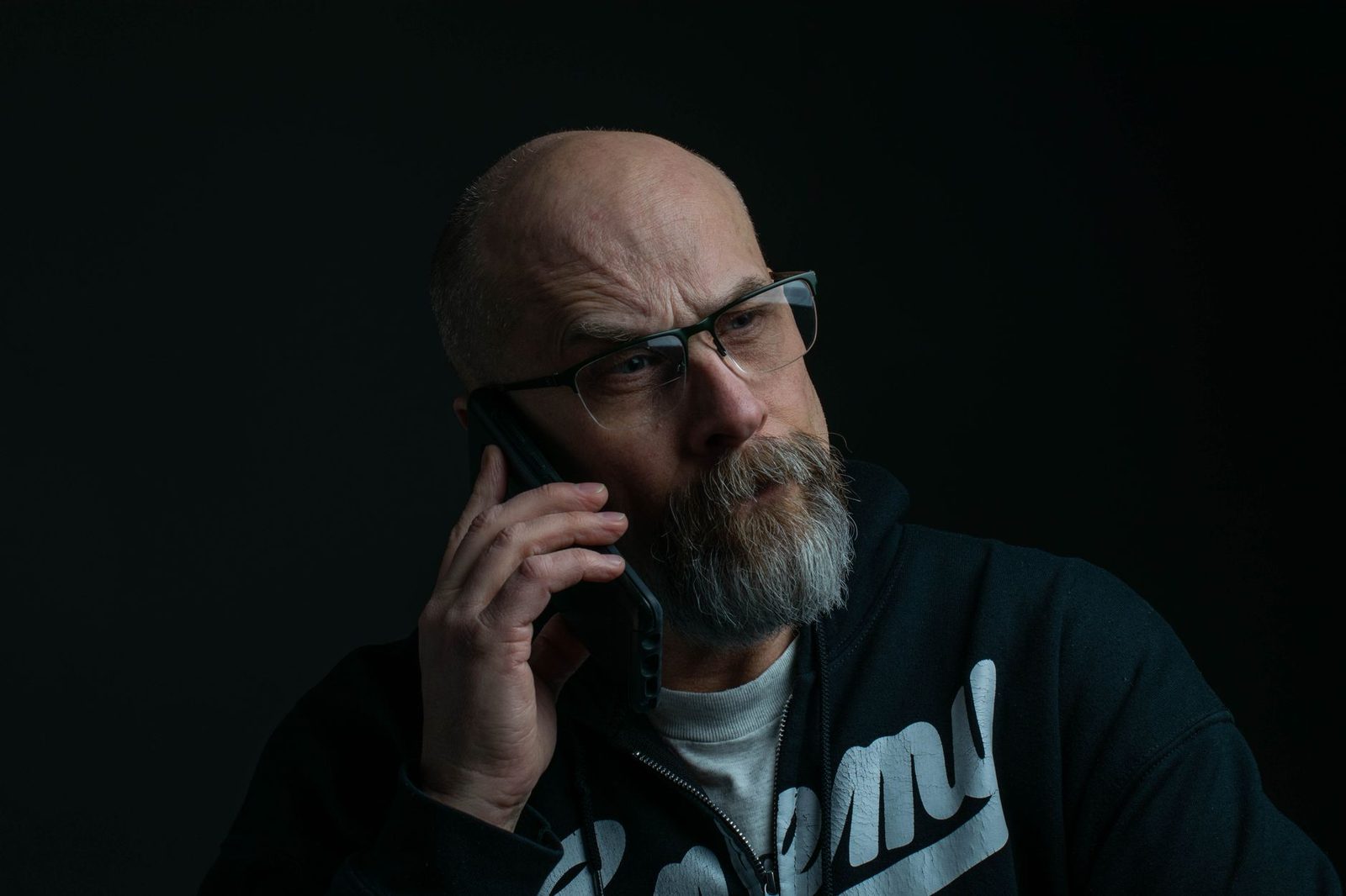 Homme barbu à lunettes, l'air inquiet, téléphonant avec un smartphone sur fond noir